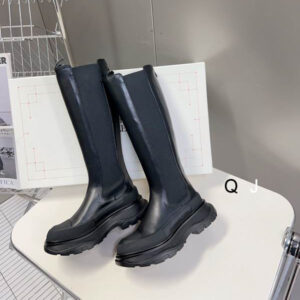 A pair of Alexander McQueen inspired black leather knee-high Chelsea boots with elastic side panels, a rounded toe, and an exaggerated, chunky black rubber tread sole.