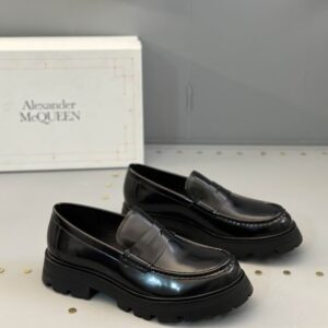 A pair of Alexander McQueen inspired black patent leather penny loafers featuring a classic strap and a very chunky, rugged black sole, displayed on a retail shelf.