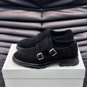 A pair of Alexander McQueen inspired black suede double monk strap shoes featuring silver buckles and a chunky black lug sole, displayed on a black leather surface next to the shoe box.