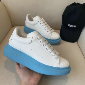 white leather Alexander McQueen Oversized Sneakers with a thick, vivid sky blue rubber sole, white laces, and Alexander McQueen branding on the tongue and heel tab, resting on a white chair.
