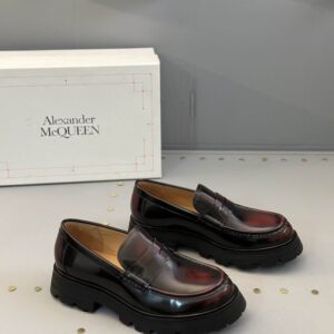 A pair of Alexander McQueen inspired oxblood/burgundy patent leather penny loafers featuring a classic saddle and a very chunky, black lug sole, displayed on a retail surface.