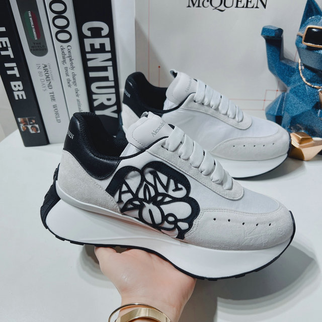 Pair of white Alexander McQueen runner sneakers with suede and textile panels, a prominent black logo on the side, and a thick, sculpted white and black rubber sole.
