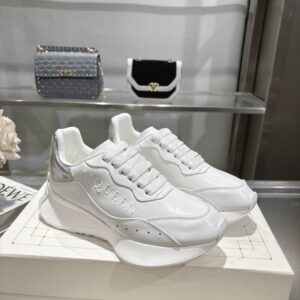 Pair of White Alexander McQueen-style runner sneakers with a large sculpted white sole and a bright Metallic Silver heel tab, displayed on a white box in a retail setting.