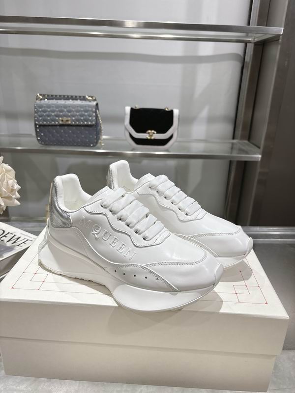 Pair of White Alexander McQueen-style runner sneakers with a large sculpted white sole and a bright Metallic Silver heel tab, displayed on a white box in a retail setting.
