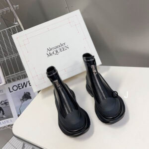 Black Alexander McQueen Tread Slick combat boots with a center zipper, thick black rubber sole with deep tread, and black leather upper, displayed on a table.