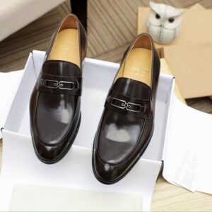 Pair of highly polished dark brown leather Berluti loafers with a silver metal buckle detail on the strap, a stacked low heel, displayed on a wooden surface.