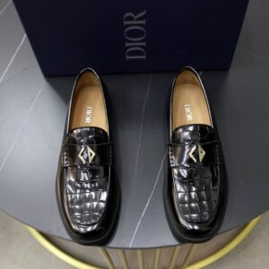 A pair of black crocodile-embossed Dior men's loafers with a gold-tone geometric buckle, displayed on a dark grey round table with a blue Dior shoe box in the background.