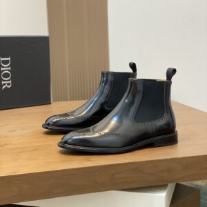 A top-down view of a pair of new, black patent leather Dior men's Chelsea boots featuring traditional brogue wingtip and medallion perforations on the toe. The boots have elastic side panels and a pull tab.