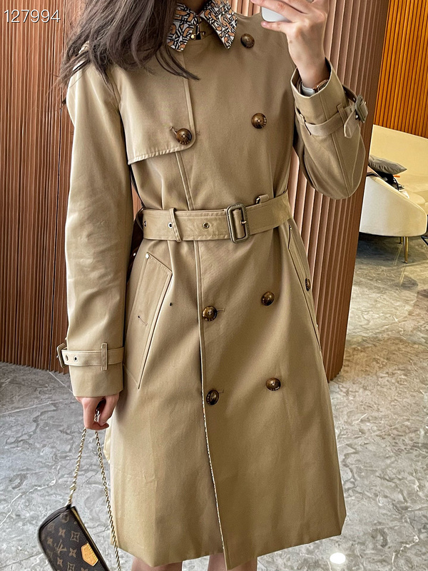 A honey-colored Burberry trench coat with a colorful TB monogram pattern on the collar, displayed on a gold rack and worn by a model.