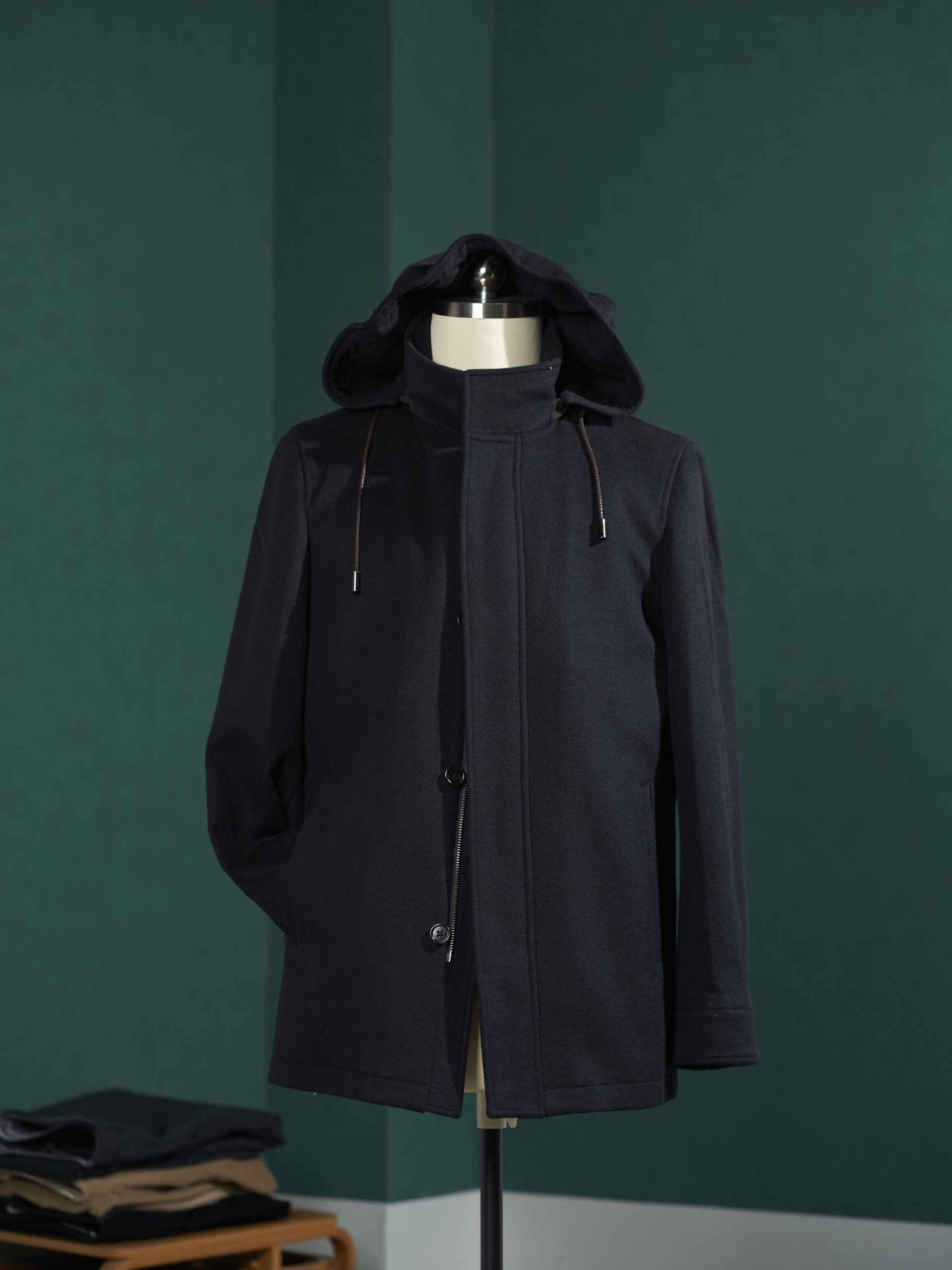 A close-up of a black wool safari jacket with four front pockets displayed on a wooden hanger against a dark green backdrop.