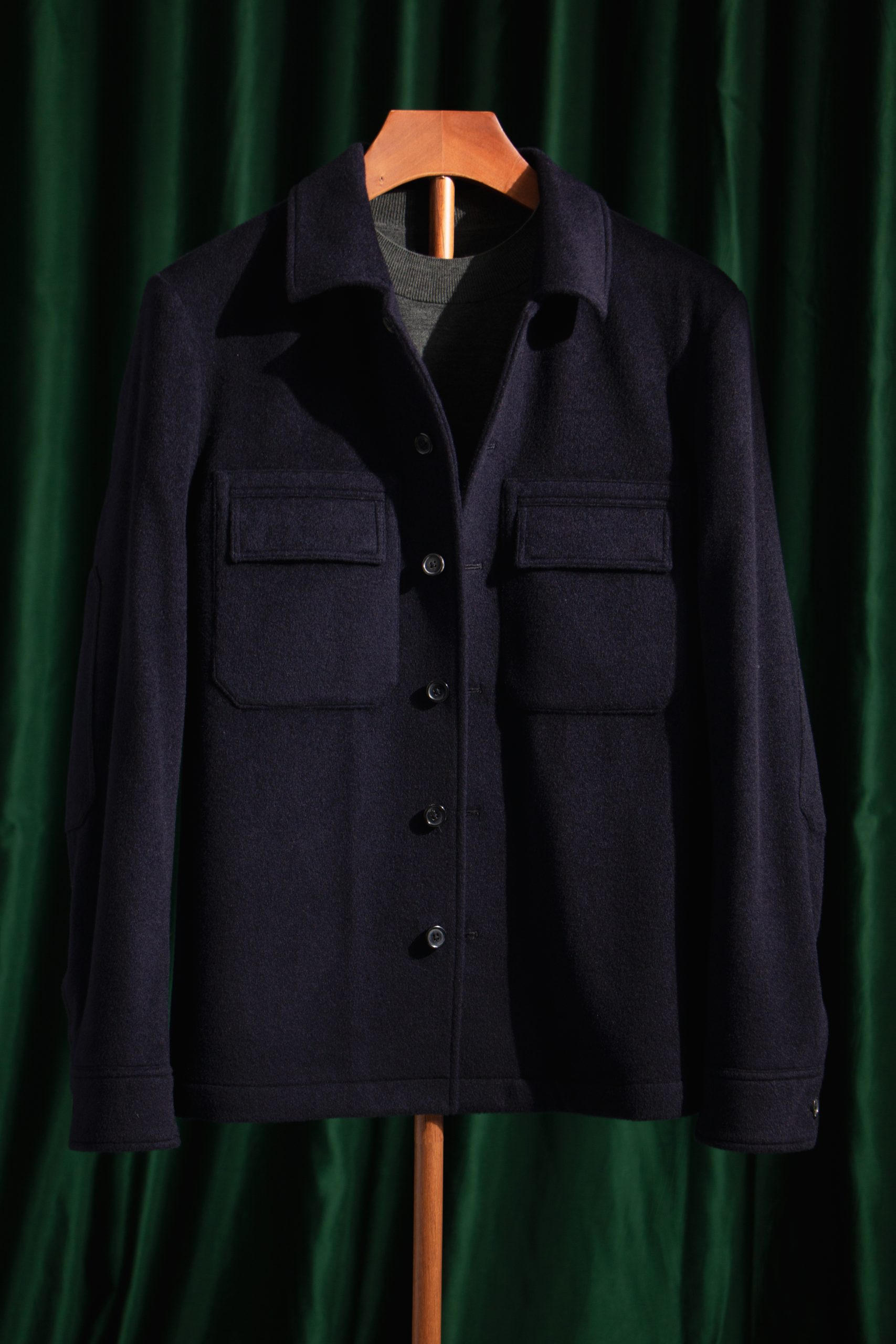 A dark navy wool safari jacket with two large chest pockets displayed on a wooden hanger against a forest green curtain.