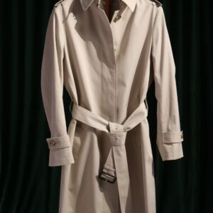 A midi-length, single-breasted beige cotton trench coat featuring an unexposed button front, back storm flaps, and a belted waist with leather buckles.