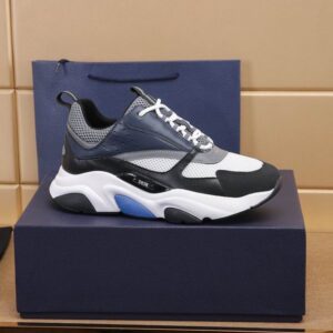 A side profile view of a DIOR B22 sneaker in navy blue and white mesh, displayed on a luxury navy blue box with a matching shopping bag in the background.