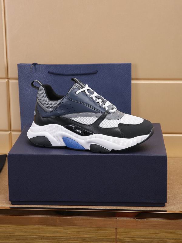 A side profile view of a DIOR B22 sneaker in navy blue and white mesh, displayed on a luxury navy blue box with a matching shopping bag in the background.