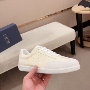 A hand holding a Dior B01 low-top sneaker featuring cream-colored leather embossed with the Dior monogram and a white suede toe cap. The sneaker has white laces, a black CD logo on the tongue, and a sleek white rubber sole, shown in a luxury showroom setting.