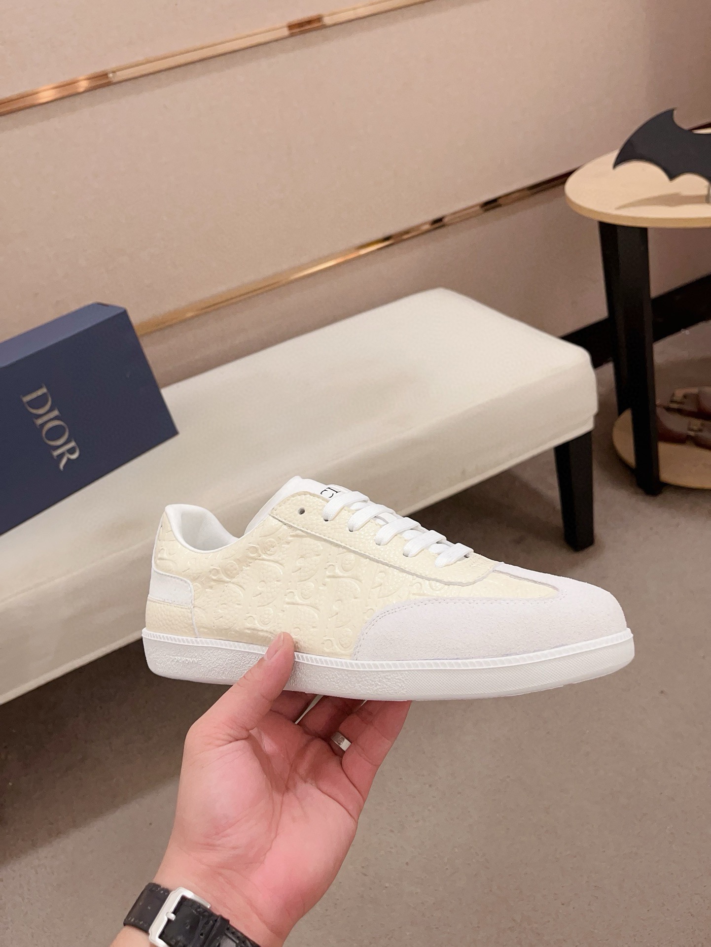 A hand holding a Dior B01 low-top sneaker featuring cream-colored leather embossed with the Dior monogram and a white suede toe cap. The sneaker has white laces, a black CD logo on the tongue, and a sleek white rubber sole, shown in a luxury showroom setting.