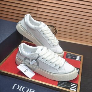 A pair of white and grey Dior B27 low-top sneakers resting on a brown textured surface next to a blue luxury Dior shoe box.