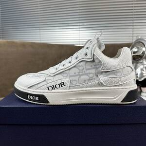 white and grey Dior B27 low-top sneakers resting on a brown textured surface next to a blue luxury Dior shoe box.