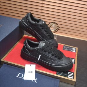 A pair of all-black Dior B33 low-top sneakers held in a person's hand. The shoes feature embossed Dior Oblique leather, thick black laces, and a chunky black rubber sole. They are shown against a backdrop of a blue Dior box and a red display tray.
