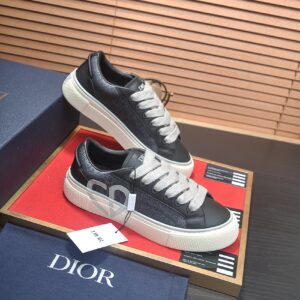 A pair of low-top Dior B33 sneakers featuring black and grey Oblique monogram fabric, black leather trim, and thick light grey laces. The side of the shoe displays a large white CD heart logo, set against a cream-colored chunky rubber sole. The shoes are displayed on a red surface with a Dior box and dust bag in the backgroun
