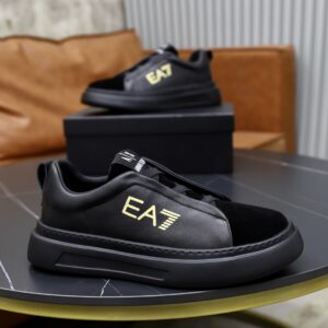 Black leather and suede chunky slip-on sneakers with a thick black sole and a metallic gold 'EA7' logo on the side, displayed on a black box.