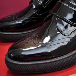 Side view of a glossy black patent leather wingtip sneaker with a velcro strap and a thick, textured black rubber sole resting on a red surface.