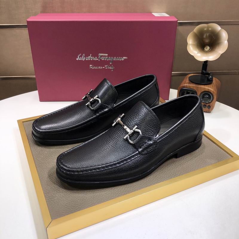 Pair of black pebble-grain leather loafers with silver bit hardware displayed on a gold tray with a magenta shoebox in the background.