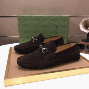 A pair of dark brown suede driving loafers with silver horsebit hardware held in a hand and displayed on a tray with a green patterned box.