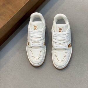 Pair of white and gold LV Trainer inspired low-top sneakers with a gum sole shown at a slight angle on a grey floor.