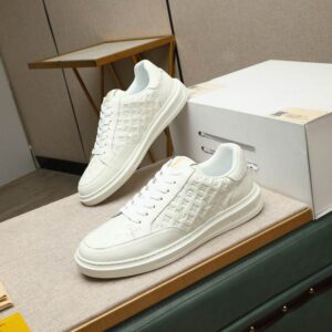 Pair of all-white low-top sneakers with an embossed monogram-inspired pattern, white laces, and a white rubber sole displayed on a cushioned table.