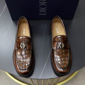 Pair of brown croc-embossed leather loafers with a gold monogram CD-style detail on the vamp and a black rubber sole, displayed on a small table.