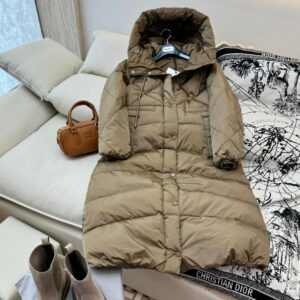 A long-length khaki brown puffer jacket with a large hood, featuring diamond-shaped quilting on the top and horizontal quilting on the bottom, shown laid out on a white sofa.