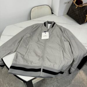Light grey Moncler lightweight bomber jacket with black and white striped rib knit trims on the collar, cuffs, and hem, lying on a white marble table next to a Dior box.
