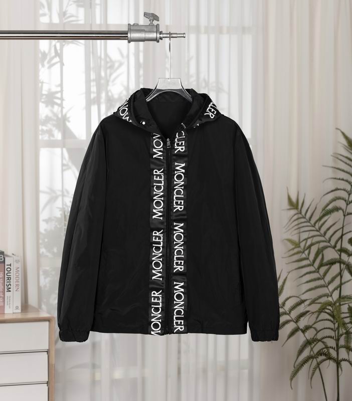 Black Moncler hooded windbreaker jacket hanging on a rail, showing a white vertical tape repeating the Moncler logo over the central zipper and around the hood edge.