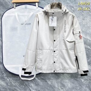 Image of a light cream/off-white technical utility jacket with a hood, black snap closures, a vertical black logo on the chest, and a utility pocket with a logo patch on the left sleeve. The jacket is presented next to a garment bag.