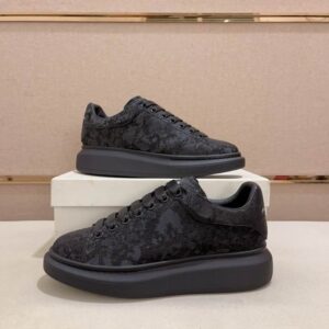 Pair of all-black, Alexander McQueen-style oversized sneakers on a white box. The shoe features a thick, sculpted black rubber sole and a black textile upper with a subtle jacquard pattern.