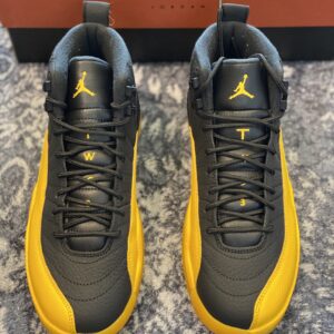 Side and top-down views of the Air Jordan 12 Retro Black University Gold sneaker on a Jordan box, featuring black leather and yellow mudguards.