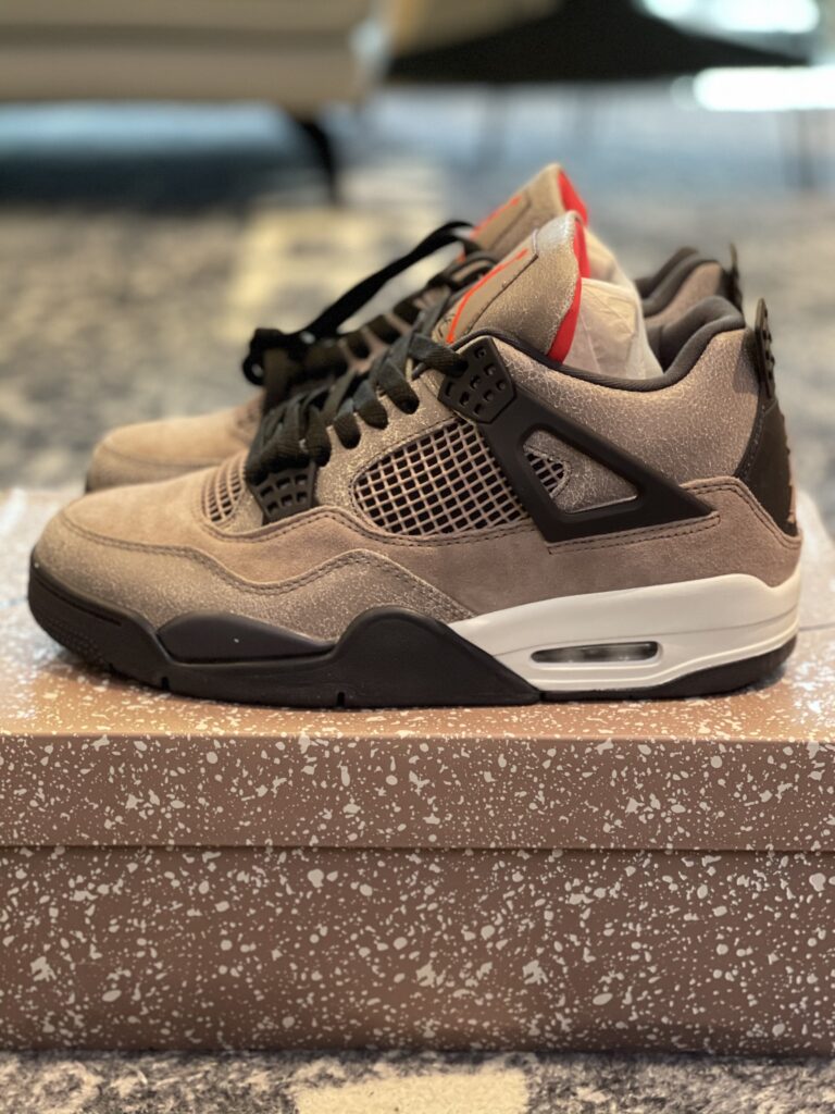 A pair of Air Jordan 4 Retro Taupe Haze Sneakers displayed on a speckled brown box, featuring taupe suede and cracked leather uppers with black accents.