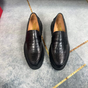 Pair of black croc-effect leather loafers with chunky black rubber soles resting on a grey floor