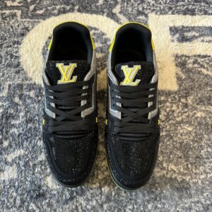 Pair of black and silver crystal-embellished LV Trainer-inspired low-top sneakers with a vibrant neon yellow sole and heel, resting on a textured gray rug.