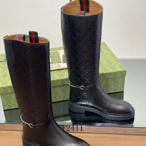 Pair of tall black leather boots with embossed GG monogram pattern on the shaft and silver horsebit chain hardware resting on a green box.