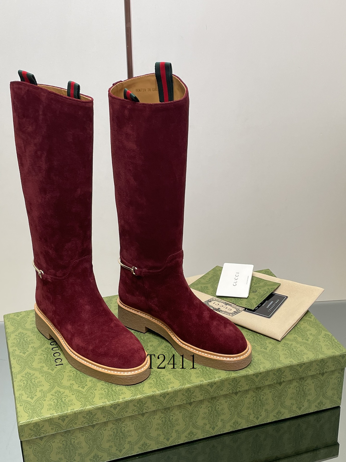 Pair of tall burgundy suede boots with gold horsebit hardware and tan rubber soles on a green logo box.