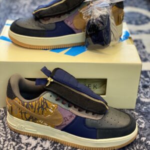 : Side profile of a LOW Travis Scott x Nike Air Force 1 inspired sneaker on a cream-colored box, showing the multicolored patchwork upper and zippered tongue.