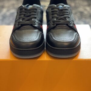 Profile view of a black grained leather LV Trainer sneaker featuring gold script branding and neon yellow accents on a luxury orange box.