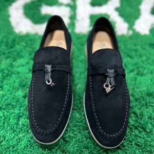 Pair of Loro Piana Summer Walk loafers in black suede featuring silver charms and a signature ivory rubber sole on a green textured surface.