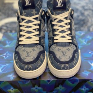 Profile view of the Louis Vuitton LV Trainer in blue monogram denim showing the side "LV" wing logo and white rubber midsole on a branded box.