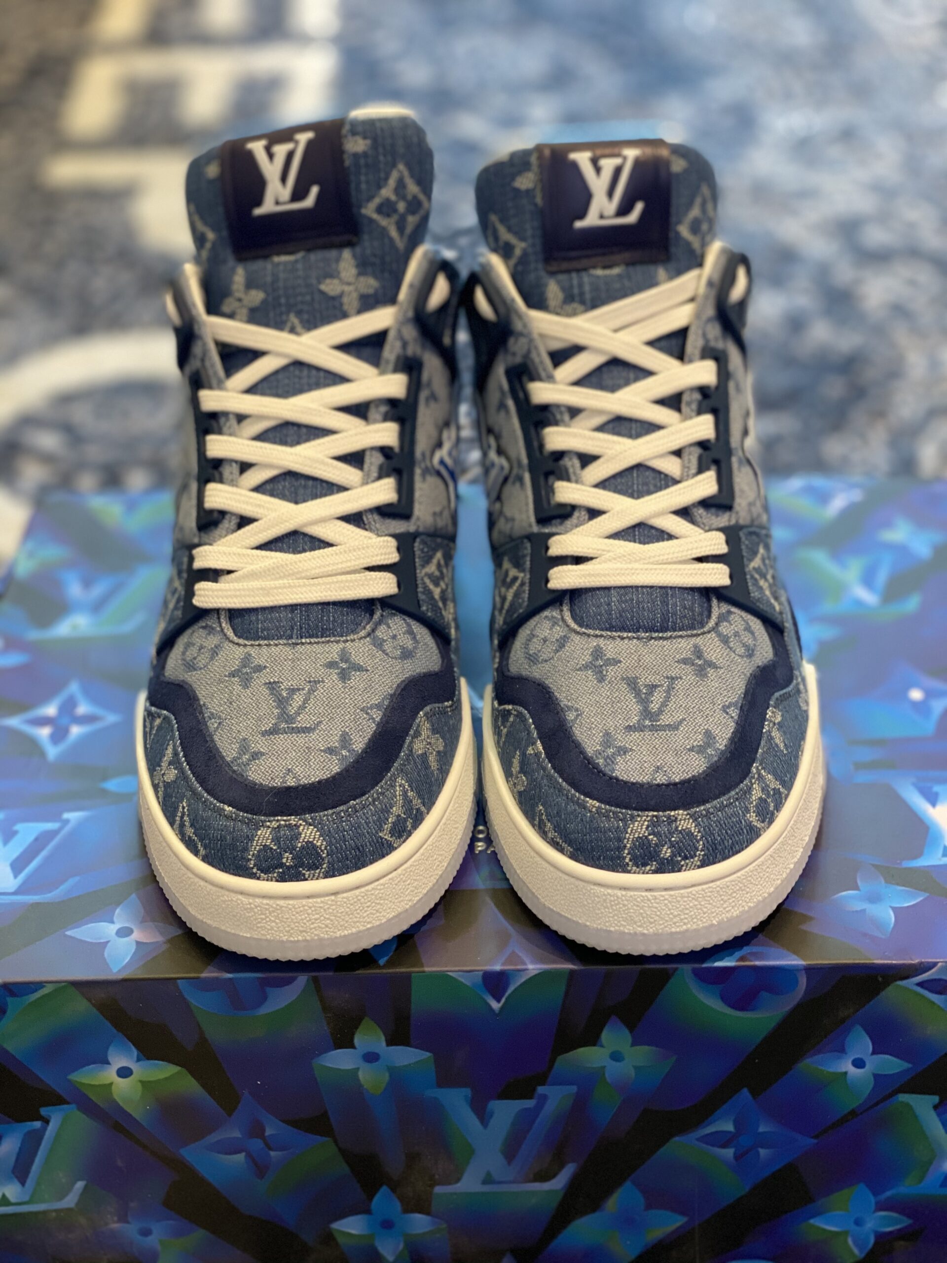 Profile view of the Louis Vuitton LV Trainer in blue monogram denim showing the side "LV" wing logo and white rubber midsole on a branded box.