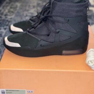 Side profile of Nike Air Fear of God 1 Triple Black sneakers sitting on an orange box, featuring a high-top design, TPU cage, and reflective toe details.