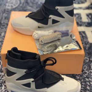 Side profile of Nike Air Fear of God 1 Triple Black sneakers sitting on an orange box, featuring a high-top design, TPU cage, and reflective toe details.