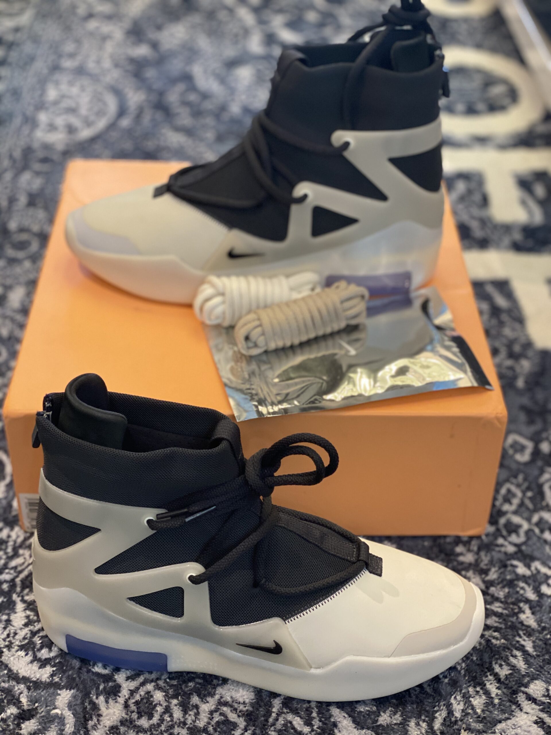Side profile of Nike Air Fear of God 1 Triple Black sneakers sitting on an orange box, featuring a high-top design, TPU cage, and reflective toe details.